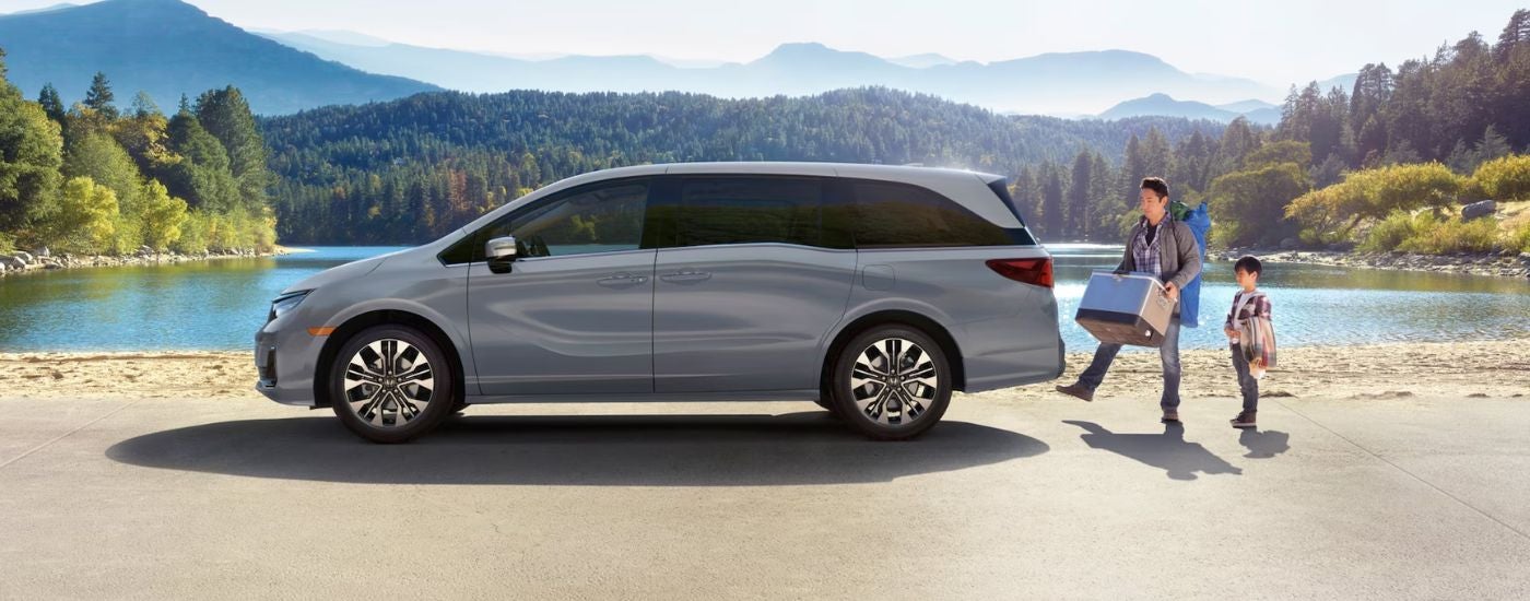 Grey 2026 Honda Odyssey parked in front of a lake, with a father loading a cooler into the cargo area.